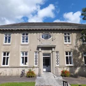 An image of Dundrum library. It is a Carnegie building in a sandy grey colour. It's a sunny day with blue skies visible behind the building. 
