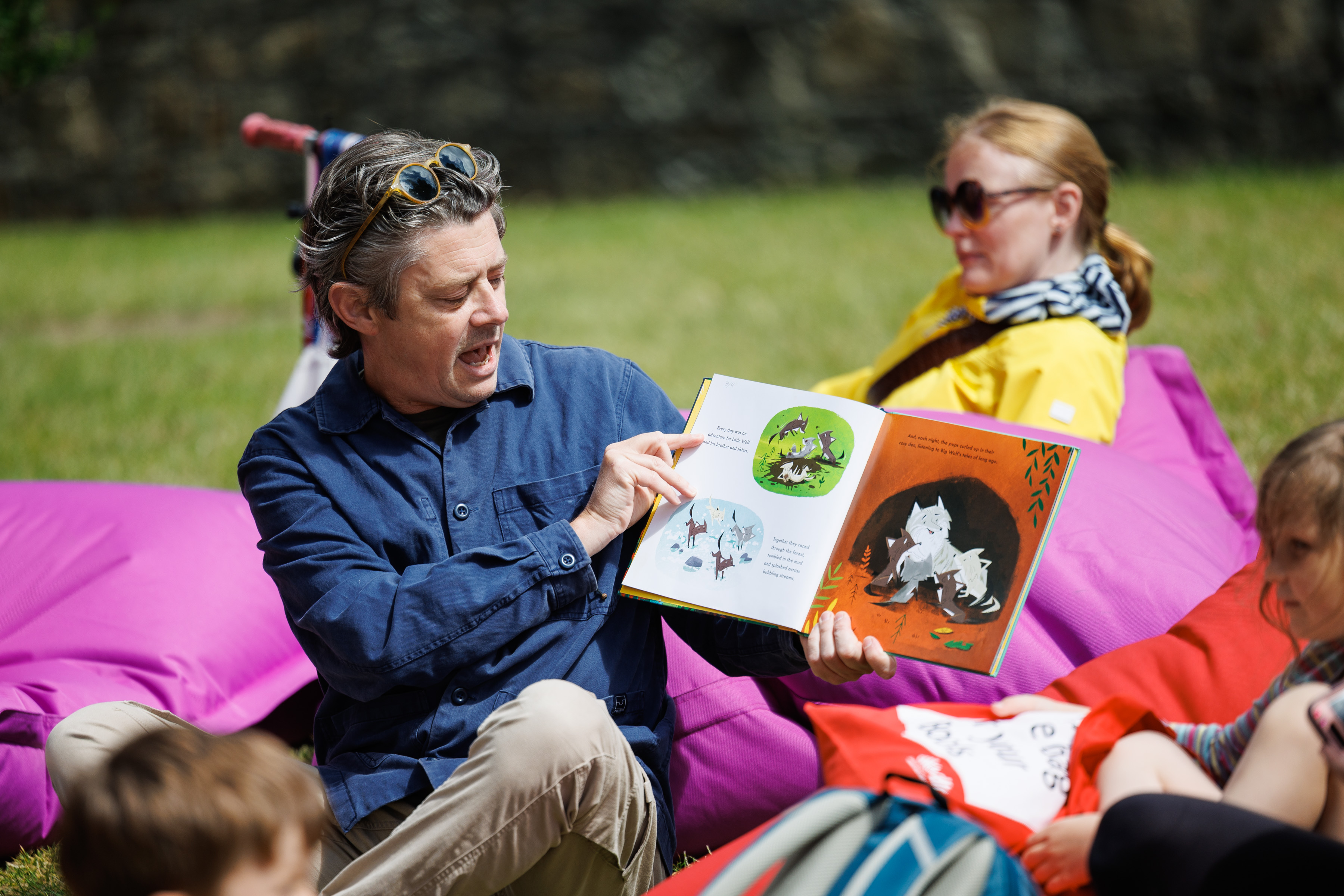 Peter Donnelly read his book Little Wolf to parents and children. He is wearing a navy shirt and sitting on grass. Parents and children sit on bean bags around him.