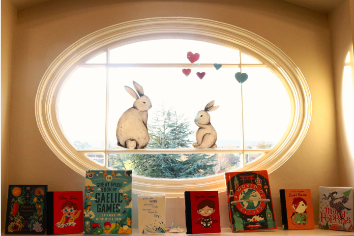 Display of children's books by a window. Cutout of rabbits with heart balloons are on the window.